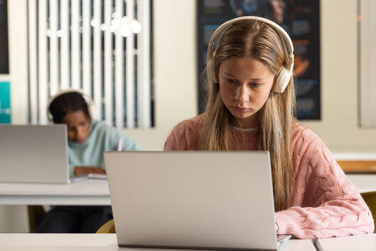 Diverse teen classmates wearing headset, female in pink sweater using laptop, male writing notebook