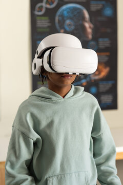 African youth male wearing white VR headset and mint hoodie standing in classroom with brain poster