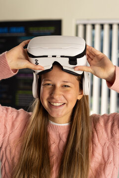 Female youth in pink sweater placing white VR headset over eyes in room by TV blinds
