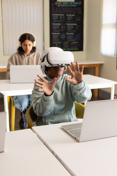 African American teenage students sitting in classroom, male wearing VR headset using silver laptop