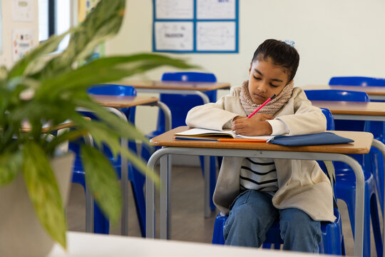 School-age African girl sitting at student desk in classroom writing in notebook with pink pencil