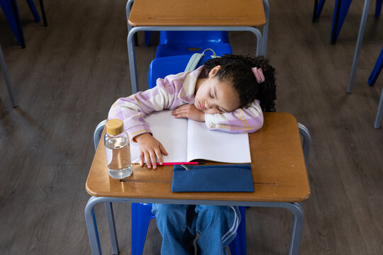 African American school-age girl resting head on open notebook at desk in classroom near red pencil
