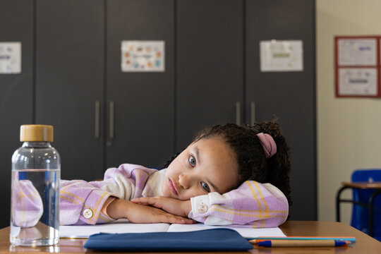 African American child student resting head on desk wearing shirt, bottle, notebook and pencil case