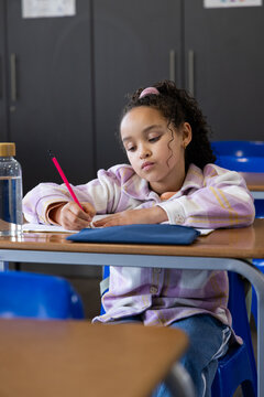 African American schoolgirl writing in classroom, holding red pencil, notebook and blue pencil case