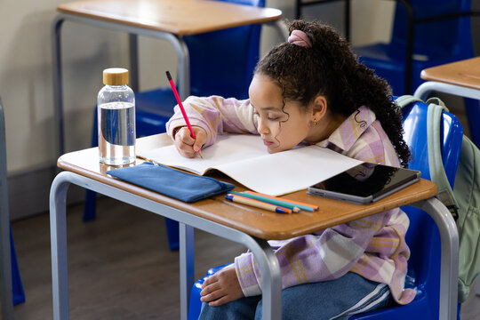 African American girl leaning, writing in notebook at school desk in plaid shirt with red pencil