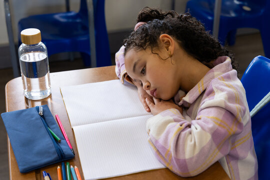 African American child sitting at wooden desk, resting head, facing open notebook, pastel plaid top