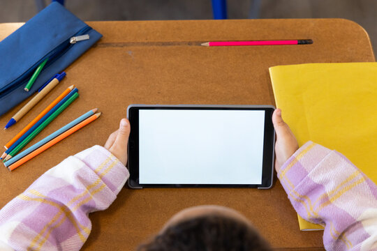 Female youth in pastel top holding tablet with blank screen on school desk with color pencils