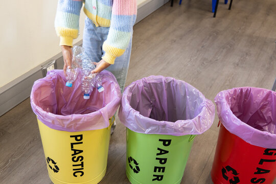 African American youth wearing pastel sweater holding PET bottles over yellow PLASTIC bin in room