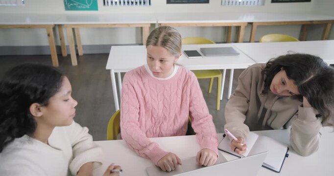 Female students at classroom table working assignment on laptop, explaining, typing, checking phone