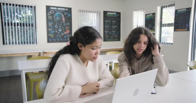 Two teenage females studying silver laptop at classroom table peer coming pointing helping homework
