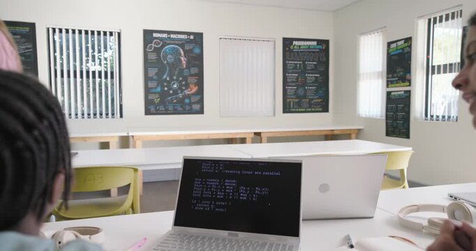 Diverse teens watching blond in pink knit walking and pointing at terminal in lab explaining code