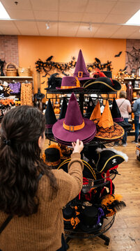 Vertical photo of a woman shopping for a Halloween costume in a retail store. Customer choosing a purple witch hat from a display rack
