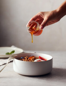 Hand pouring honey over a yogurt and granola breakfast bowl with fresh berries, healthy breakfast and clean eating concept with copy space