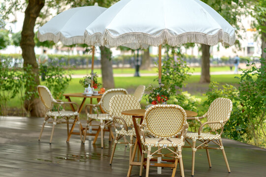 Outdoor cafe terrace with wicker chairs and wooden tables under vintage white umbrellas. Cozy summer restaurant veranda with elegant patio furniture setup in a park area.