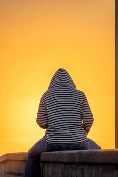 Saint-Aubin-Sur-Mer, France - 07 27 2026: A woman with a navy blue fleece is sitting with her back turned on the jetty, the beach and the sea on the sun at golden hour