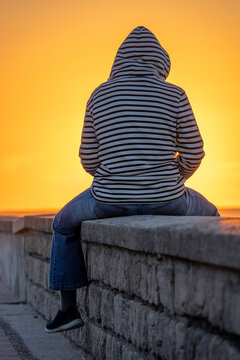 Saint-Aubin-Sur-Mer, France - 07 27 2026: A woman with a navy blue fleece is sitting with her back turned on the jetty, the beach and the sea on the sun at golden hour