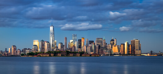 Naklejka premium Panoramic view of the Lower Manhattan skyline and Ellis Island over the New York City harbor at dusk with golden light reflecting on skyscrapers