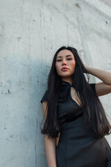 Fototapeta premium Confident Asian woman in black cheongsam-style dress against textured concrete. Long hair, powerful gaze from low angle. Modern and stylish.