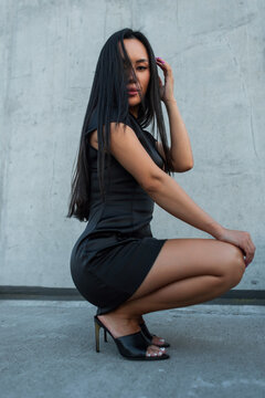 Side profile of a beautiful Asian woman crouching in a short black silk dress and high heels against a concrete wall. Pretty hot elegant beauty girl