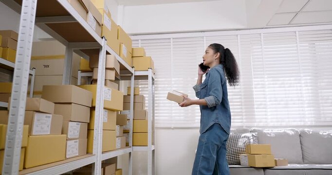 Asian woman small business owner talks on smartphone while walking through warehouse, reviews customer order status, checks parcel box placement and supports e-commerce order fulfillment process