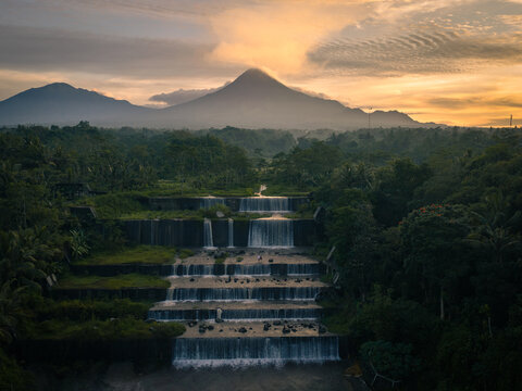 Aerial view of Grojogan Watu Purbo, a tiered waterfall cascading through lush greenery with a volcano in the background at sunset in Kecamatan Tempel, Daerah Istimewa Yogya, Indonesia.