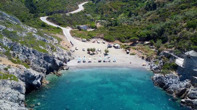 Aerial drone footage of Paralia Gialia in Alonissos, Greece, featuring turquoise waters, a secluded pebble beach and the historic traditional windmill on a sunny summer day.