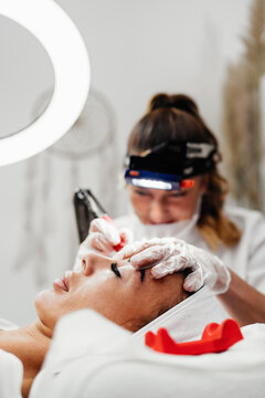 Specialist performing eyebrow microblading treatment using professional tattooing tool on client lying on treatment bed in aesthetic clinic.