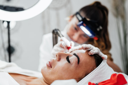 Specialist performing eyebrow microblading treatment using professional tattooing tool on client lying on treatment bed in aesthetic clinic.