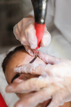 Close-up shot of a specialist applying semi-permanent makeup and pigment to a client's brow line using specialized equipment and sterile gloves during a beauty treatment.