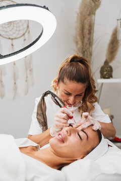 Specialist performing eyebrow microblading treatment using professional tattooing tool on client lying on treatment bed in aesthetic clinic.