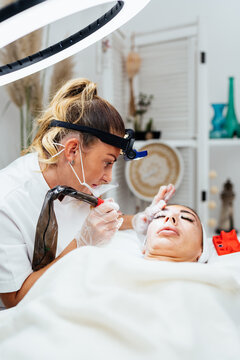 Specialist performing eyebrow microblading treatment using professional tattooing tool on client lying on treatment bed in aesthetic clinic.
