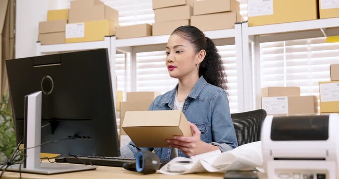 Asian woman entrepreneur holding shipping box while typing customer order data, checking inventory system, managing online shopping parcel box preparation, coordinating e-commerce delivery workflow