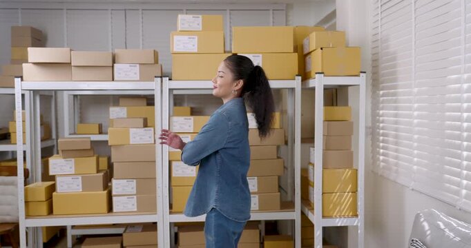 Asian woman small business owner stands confidently with crossed arms in warehouse surrounded by parcel boxes representing successful e-commerce business operation