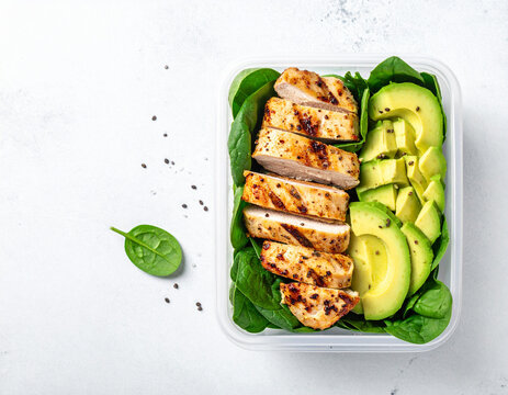 Clinical chic meal prep container showing layered grilled chicken breast strips with spinach and fresh avocado chunks.