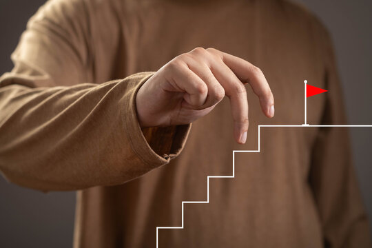 Close-up of a hand moving toward a staircase leading to a red flag, symbolizing goal achievement, career growth, business success, leadership, motivation, progress, ambition, and strategic planning.