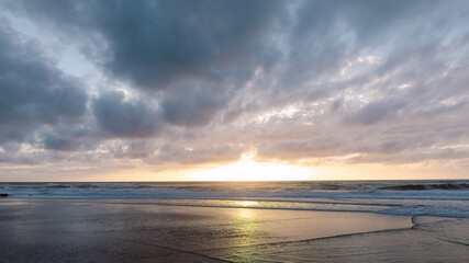 Obraz premium Golden sunset glows on the horizon over an ocean beach, with soft waves rolling onto the wet sand and dark clouds overhead