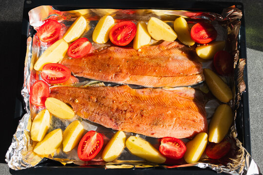 Two salmon fillets are arranged on a baking tray surrounded by sliced potatoes and tomatoes. This scene captures the preparation of a healthy meal in a kitchen setting