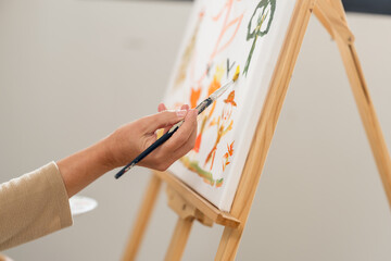 Senior woman applies orange paint with brush onto colorful canvas on wooden easel, practicing art therapy at home for creative expression, mindfulness, relaxation, wellness, and active retirement. © Ekkasit A Siam