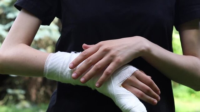 Person touches a white gauze wrap on their injured wrist during a sunny afternoon outdoors while wearing a black shirt to assist with the recovery and healing process of a recent physical injury