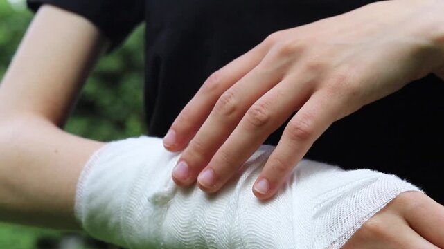 Patient wrapping a soft white medical bandage around their wrist while standing outside in a park during the day to protect and support an injured arm during the recovery and healing process