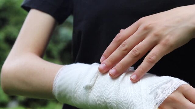 Injured person touches a white gauze bandage wrapped around their sore wrist during the day to check the security of the medical support after experiencing a painful arm injury in a park setting