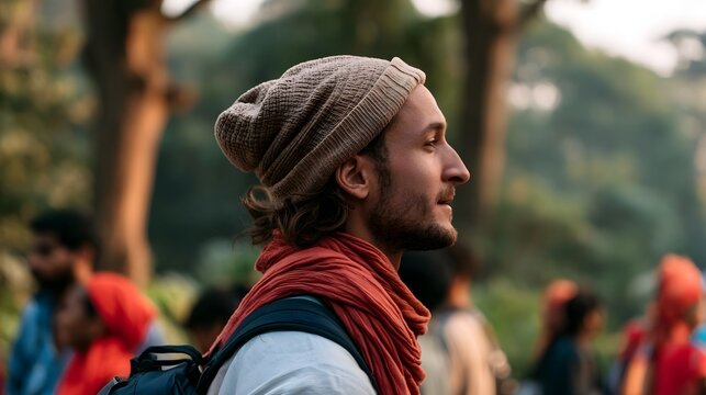 A man wearing a knit beanie and red scarf looks thoughtfully to the side while standing outdoors among blurred people