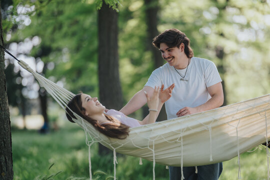 A young couple relaxes in a hammock while the man smiles and interacts with the woman. They enjoy a peaceful, sunlit moment together in a leafy park setting.