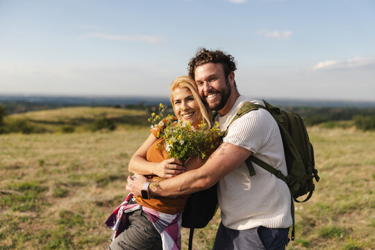 Couple Smiling While Holding Wildflowers on a Hiking Trail in a Sunny Landscape