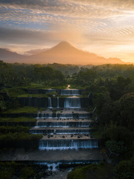 Aerial view of Grojogan Watu Purbo featuring tiered waterfalls and stone structures with a volcano in the background at golden hour in Kecamatan Tempel, Daerah Istimewa Yogya, Indonesia.