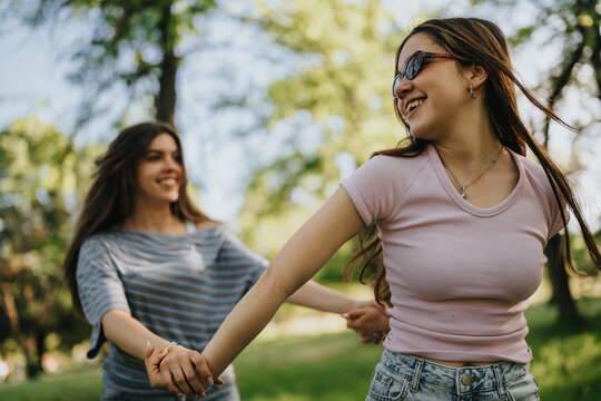 Two teen friends hold hands and smile while walking outdoors. The girls enjoy a sunny park moment together, looking happy and carefree.