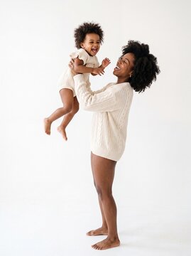Happy Black mother holding her laughing toddler baby girl in the air against a seamless white background in cozy matching knitted clothing