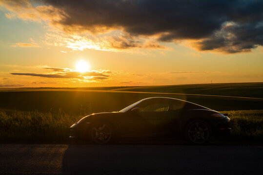 Porsche Cayman, silhouette of streamlined lines against the backdrop of a picturesque sunset over the waves of fields.