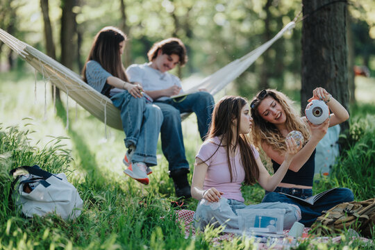 Teen friends relax and share objects while sitting on a blanket and hammock in a grassy park. A group of young people enjoy a casual outdoor hangout together in nature.