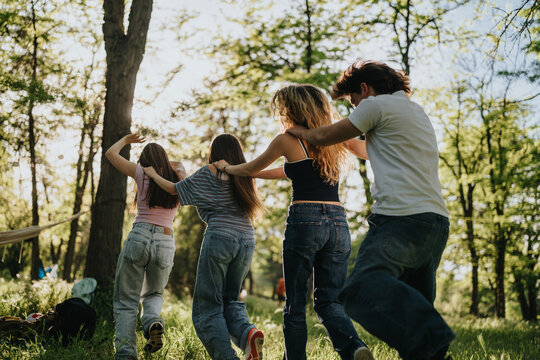 Four teen friends walk together with arms linked through a sunlit forest clearing. They move in a line across grassy woods, sharing a casual outdoor moment.